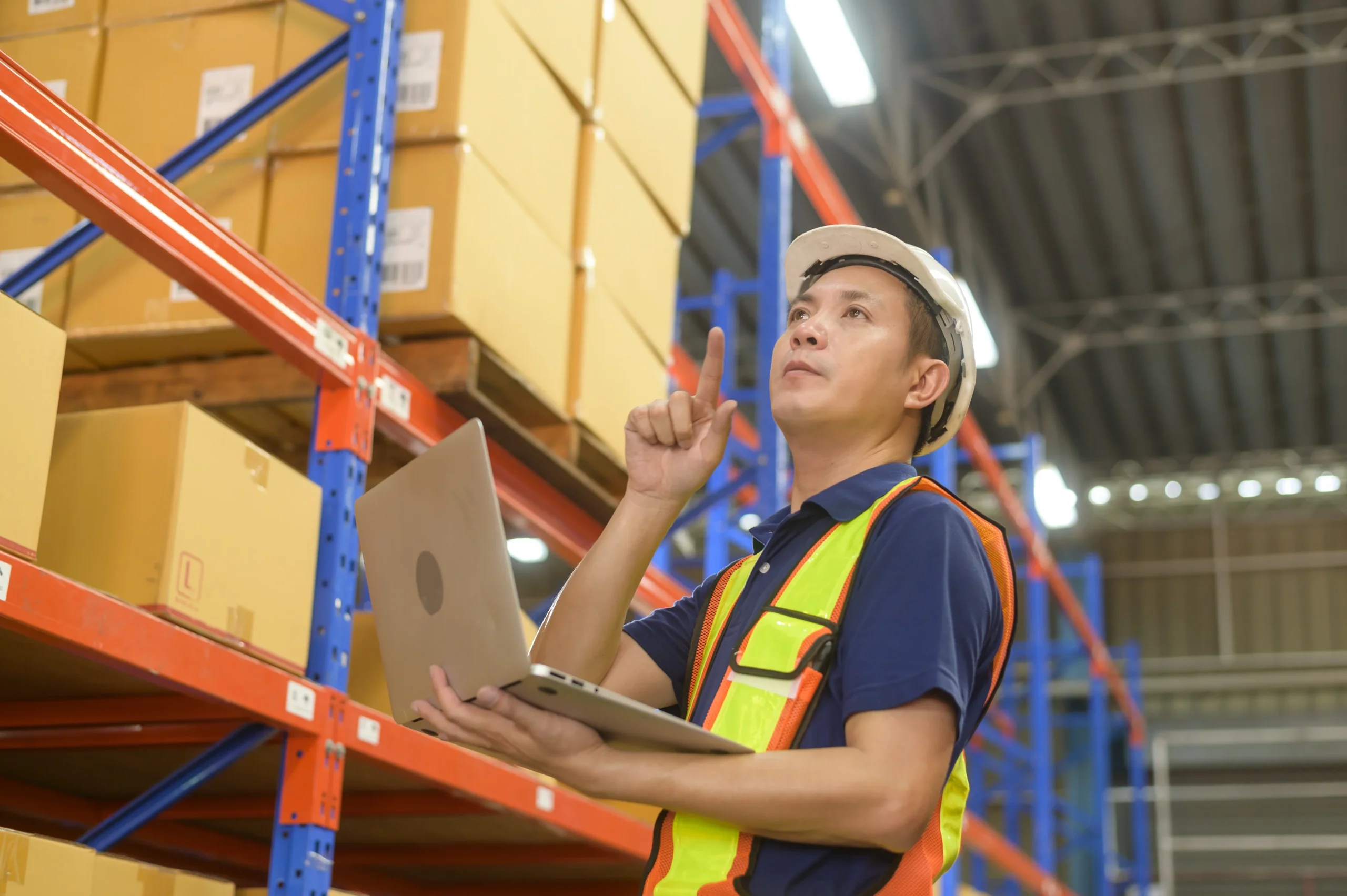 Warehouse manager wearing a safety helmet and reflective vest, using a laptop to assess inventory in a warehouse, demonstrating warehouse management system evaluation.