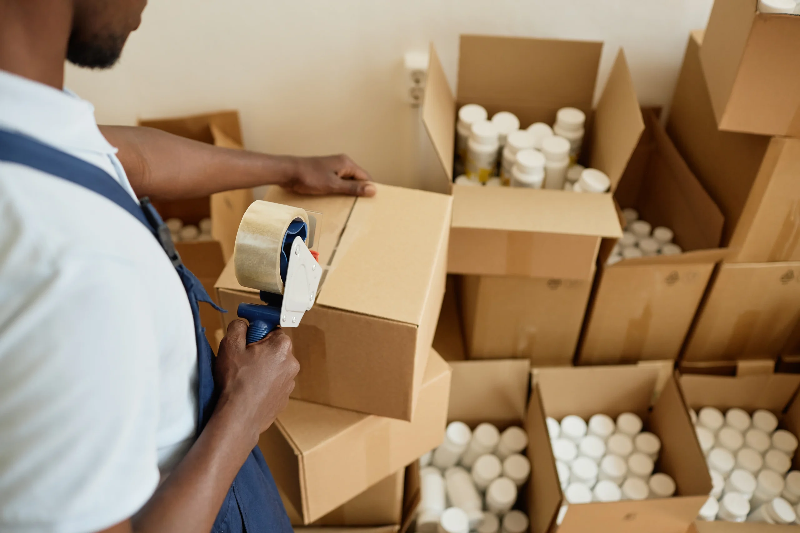 Pick and pack worker using tape to seal a cardboard box in a warehouse filled with products.