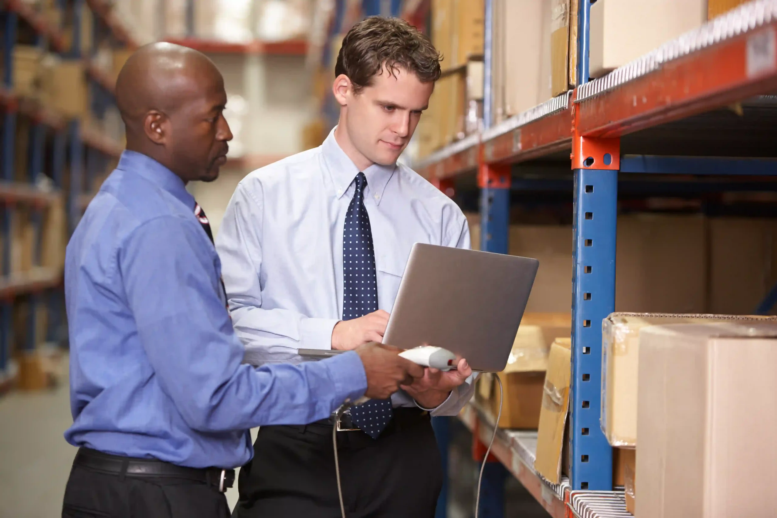 Two business professionals using a laptop to review an inventory management system questionnaire in a warehouse.