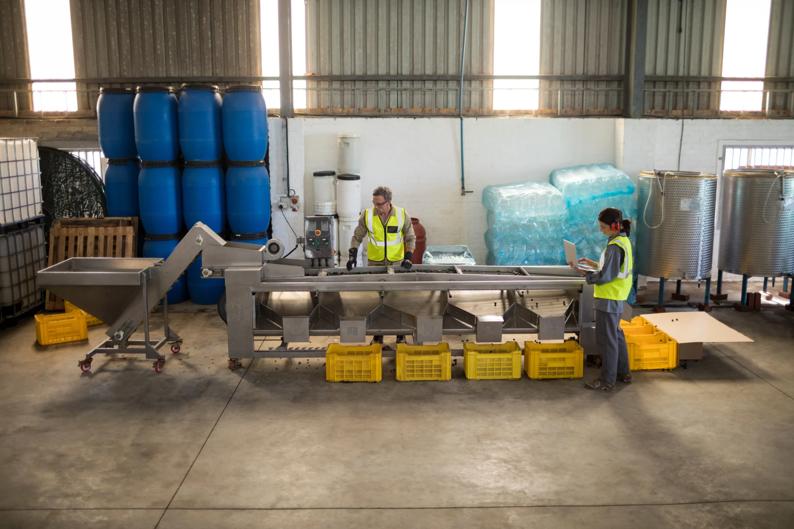 Warehouse team working near a production line with storage barrels and crates, optimizing logistics operations.