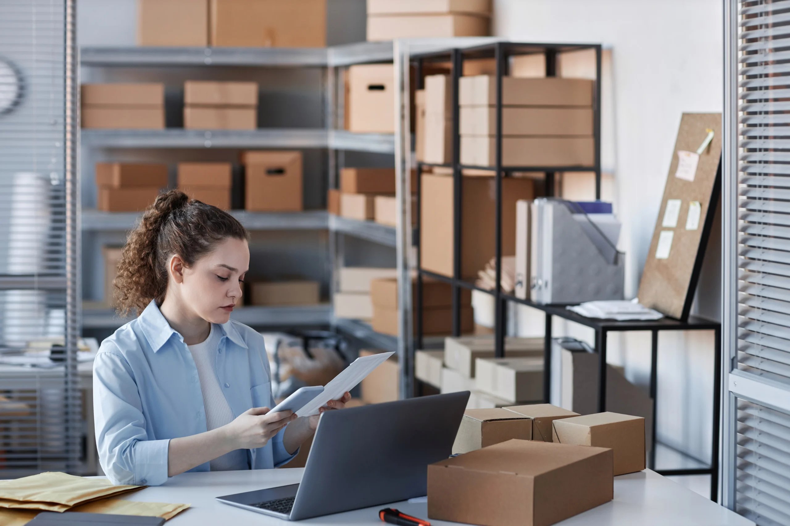 Young woman in a warehouse using a smartphone and laptop to manage inventory records with an inventory management system in Google Sheets.