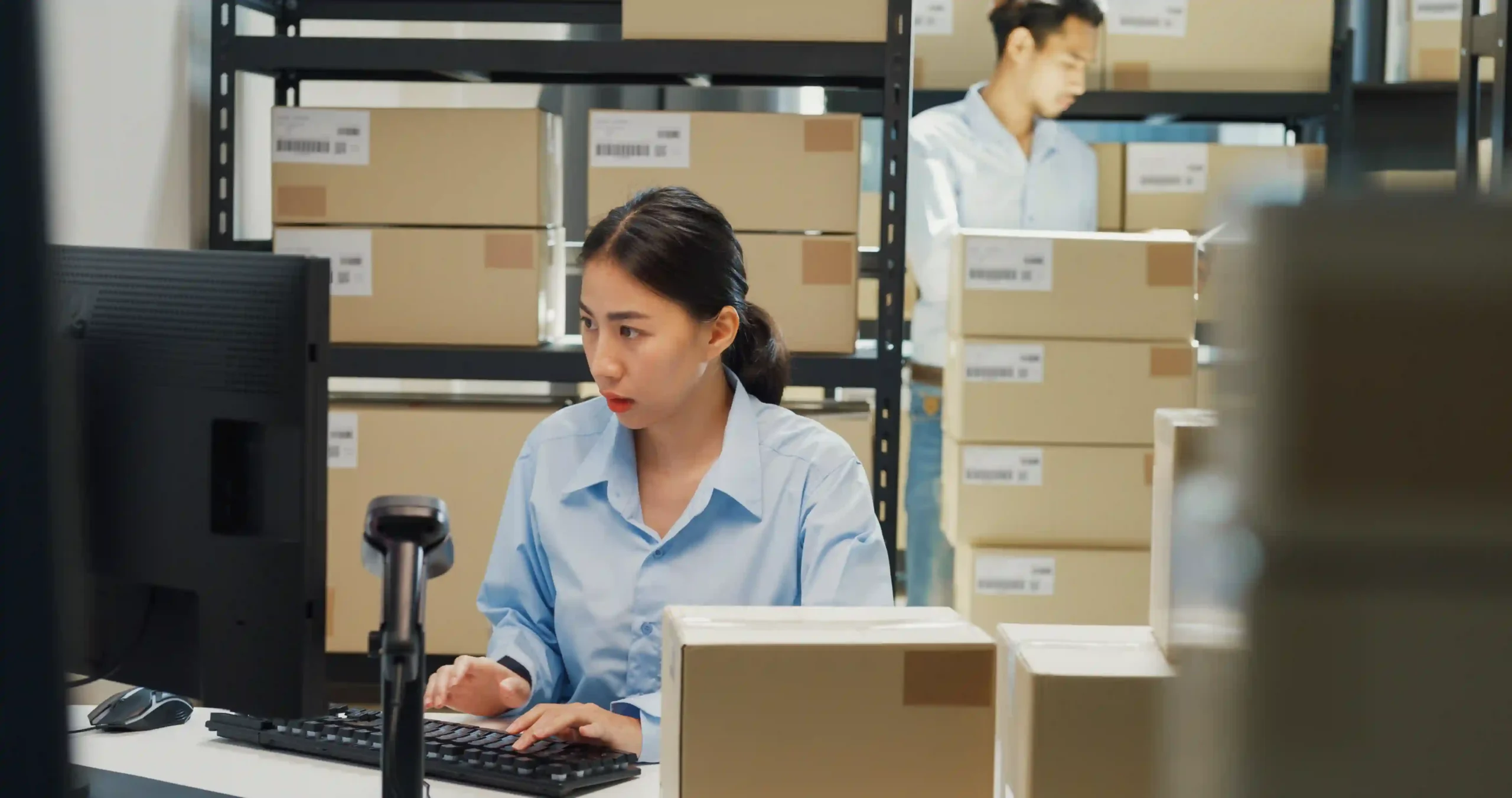 Female warehouse employee managing order processing in warehouse using desktop system and barcode scanner.
