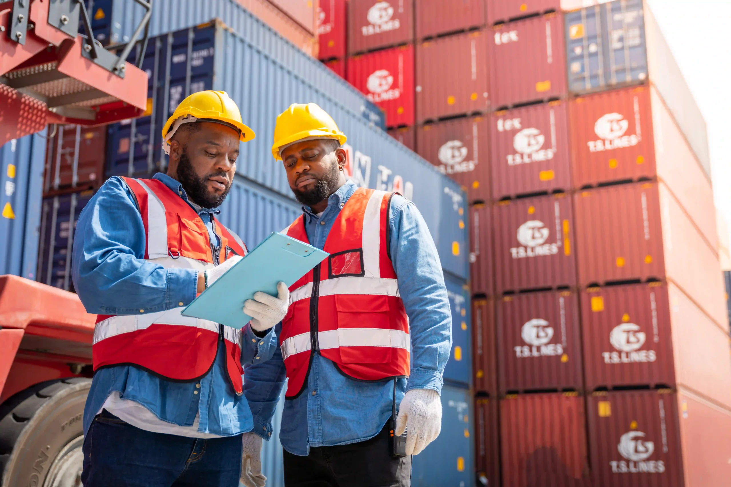 Two logistics workers reviewing shipment data on a clipboard in front of stacked containers, showcasing teamwork in shipping logistics careers.