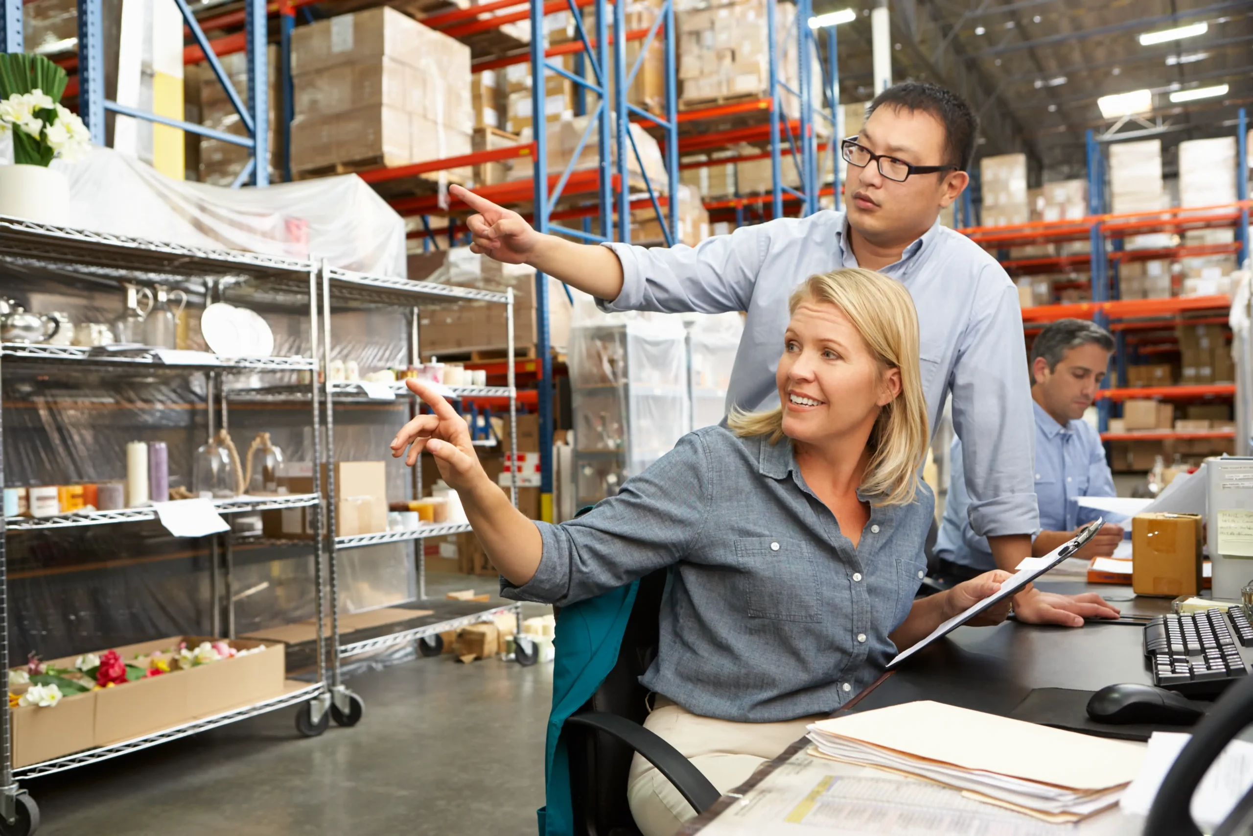 Team collaboration at a desk in a busy warehouse with inventory shelves in the background.