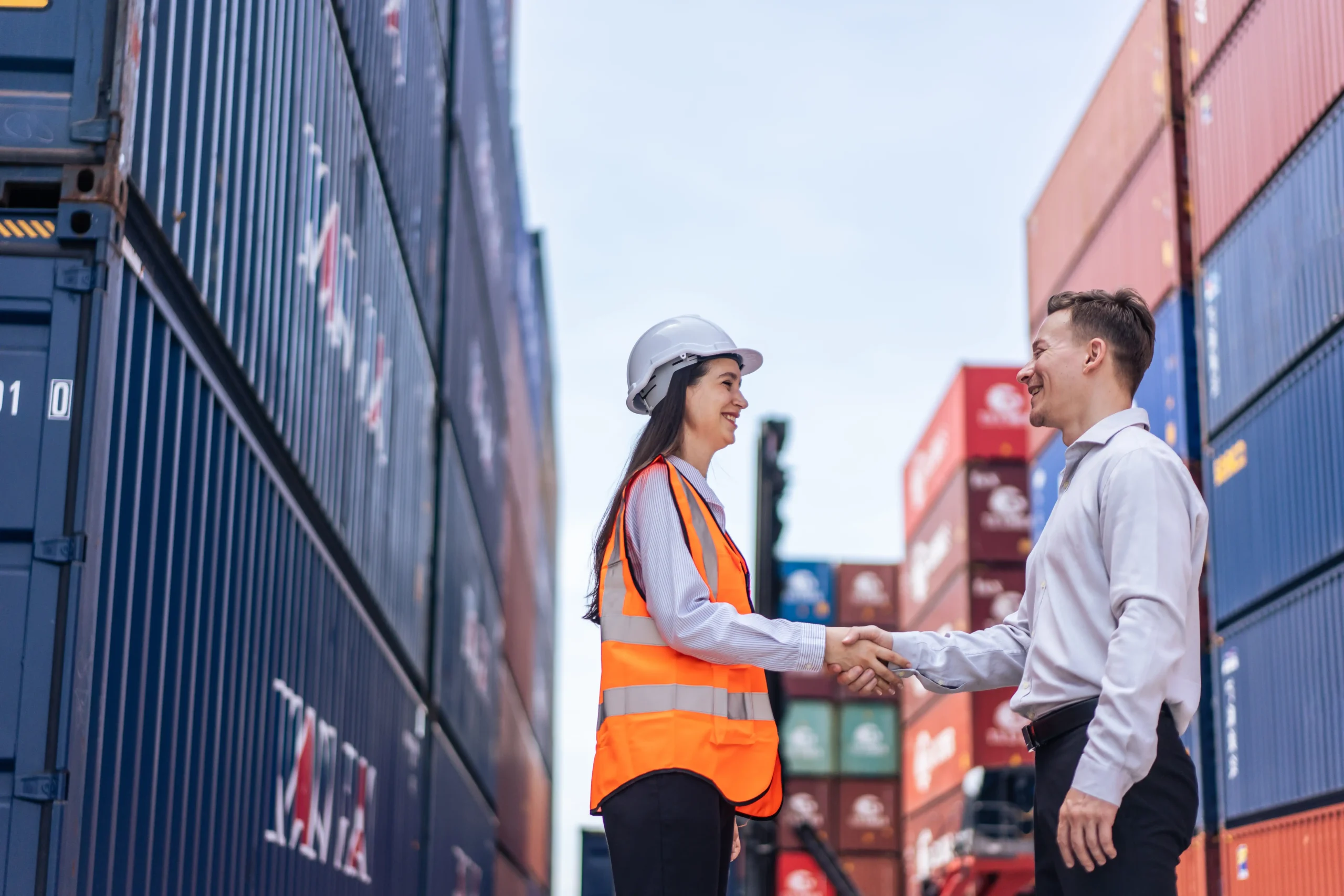 Female warehouse worker and business professional shaking hands near shipping containers, symbolizing logistics partnership.