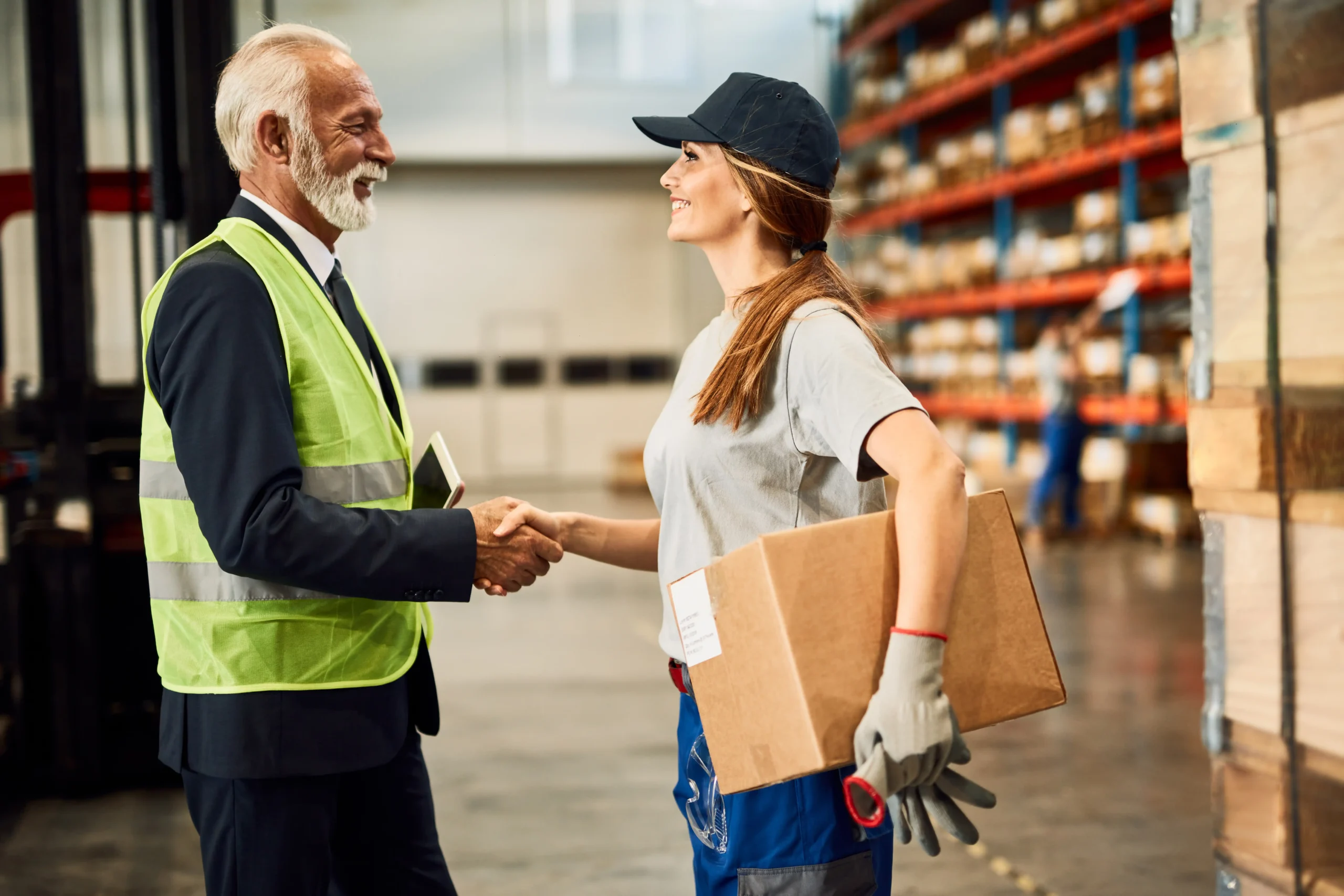 Senior businessman shaking hands with a female warehouse worker in a distribution center, symbolizing successful logistics services.