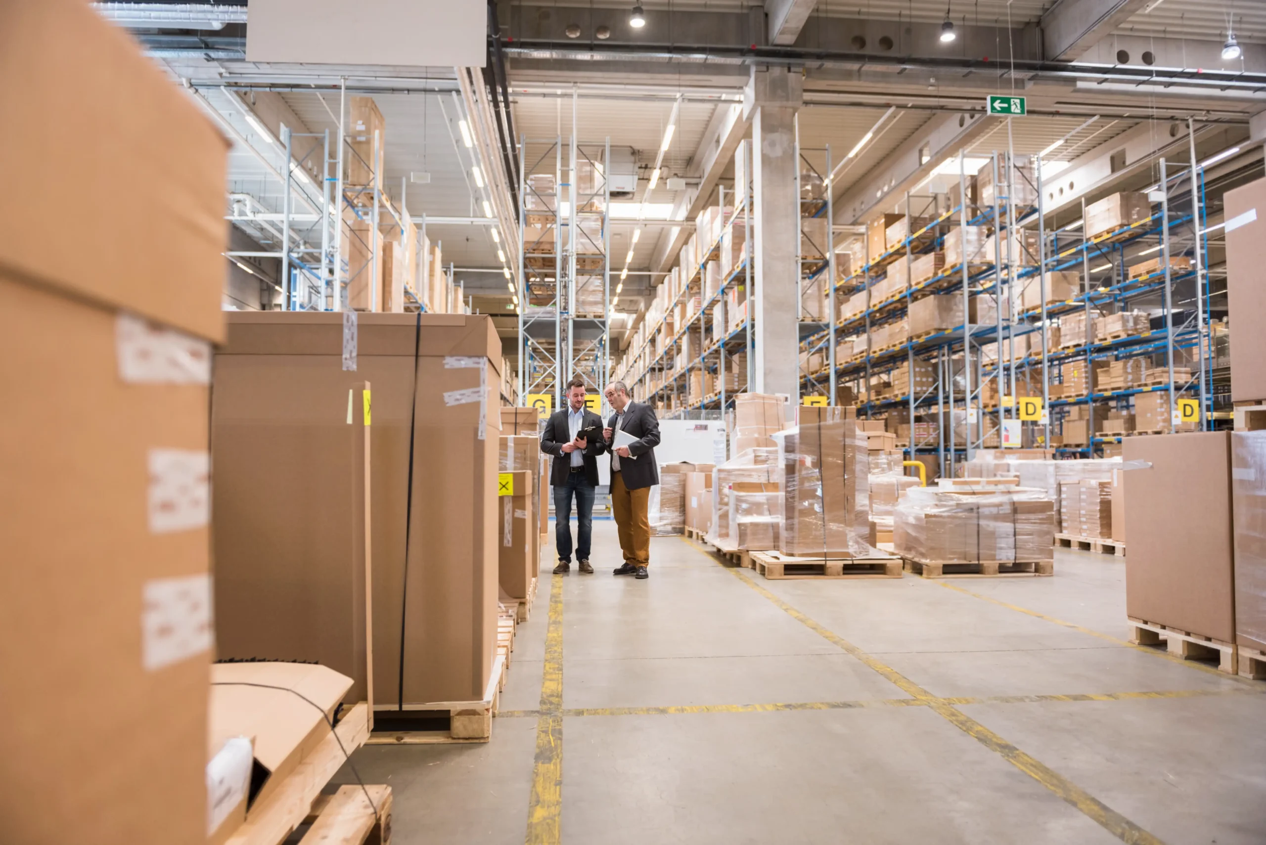 Two men discussing logistics operations in a factory warehouse surrounded by inventory.