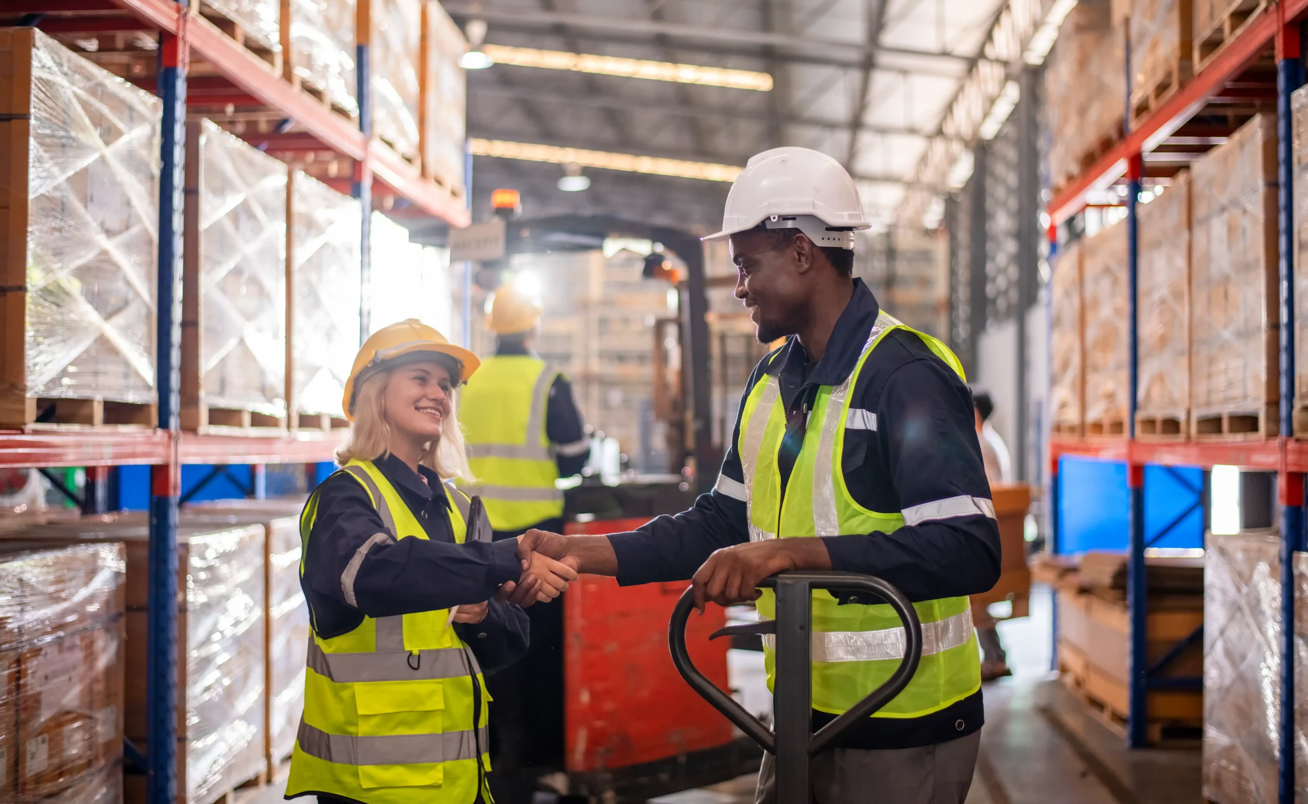 Warehouse workers in safety vests and helmets shaking hands in an industrial setting with inventory racks.