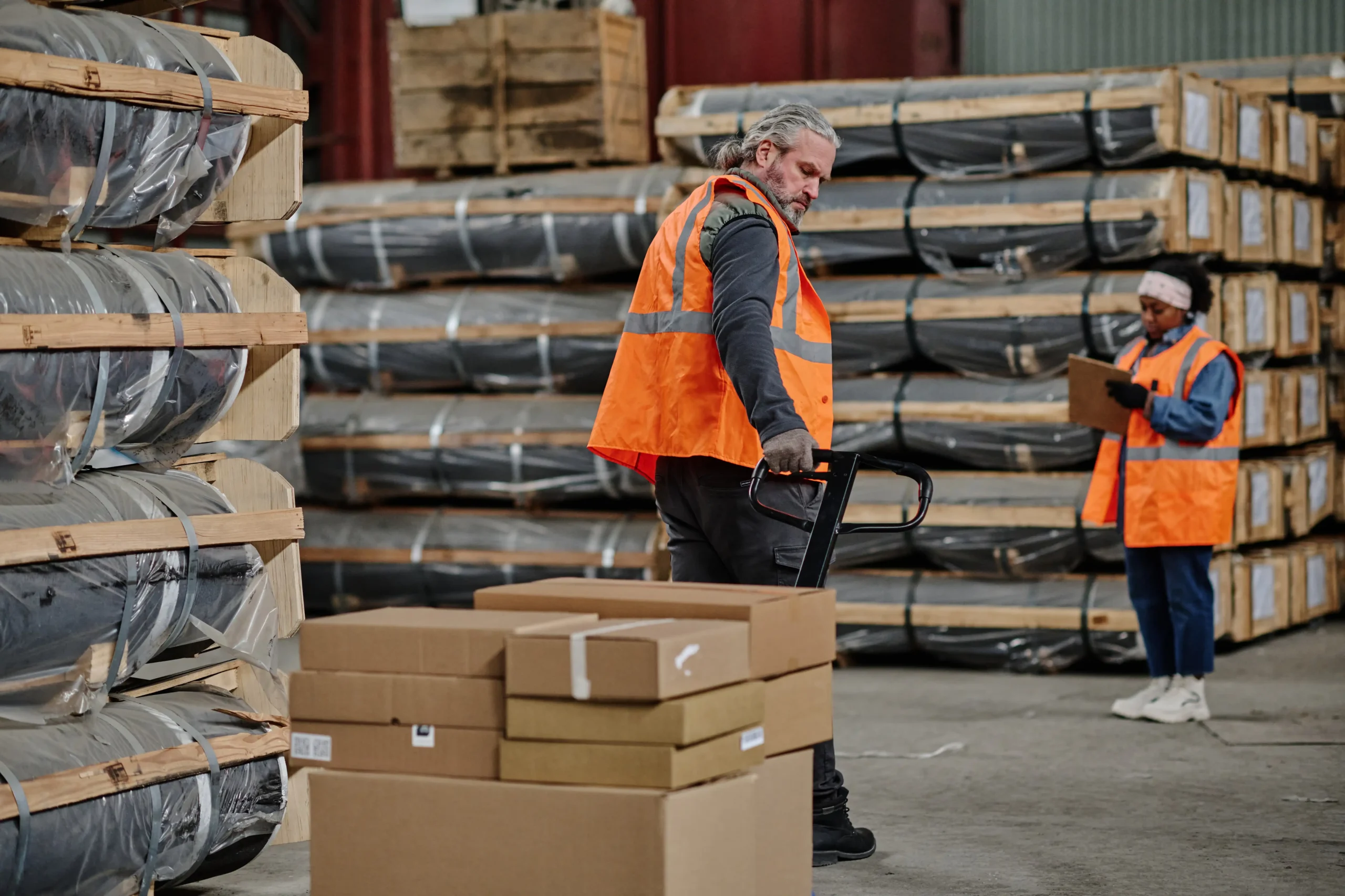 Worker operating a hand pallet truck in a warehouse while another worker inspects inventory with a clipboard.