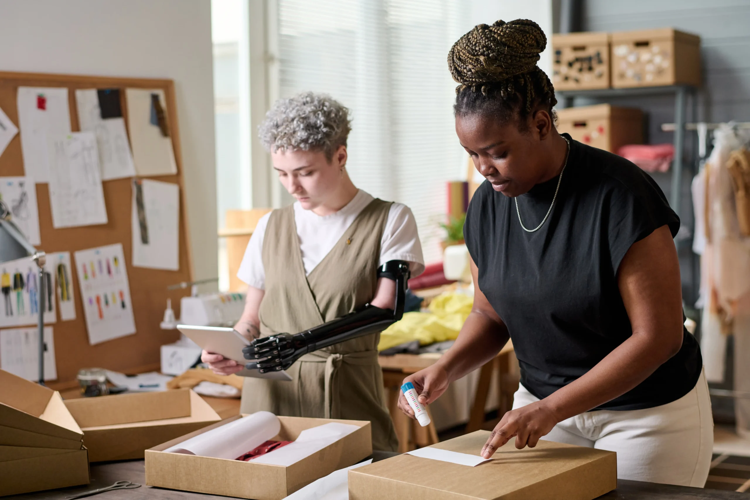 Two women packing e-commerce orders in a fulfillment workspace with one checking inventory on a tablet.