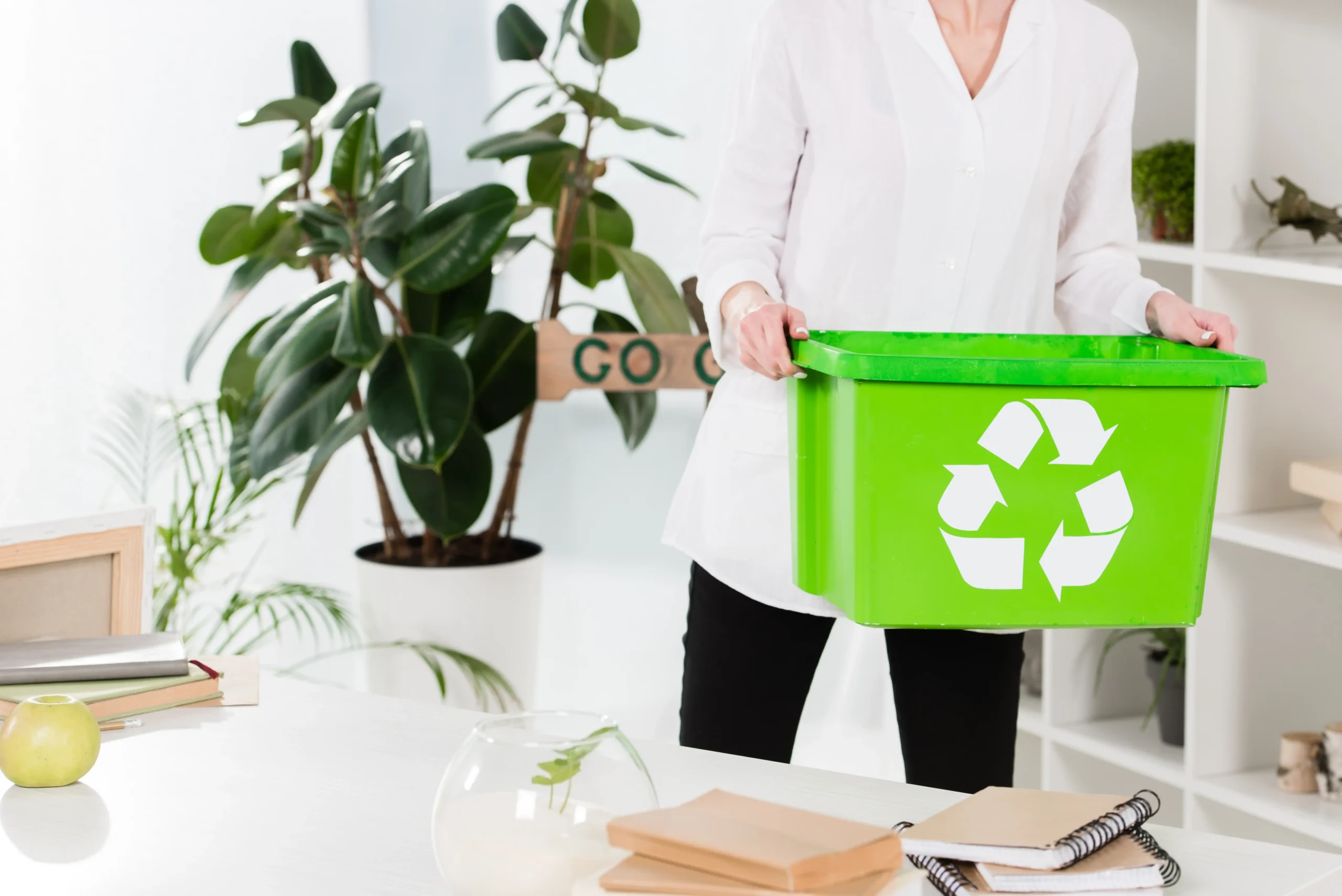 Woman holding a green recycling box in a bright office with plants promoting sustainability.