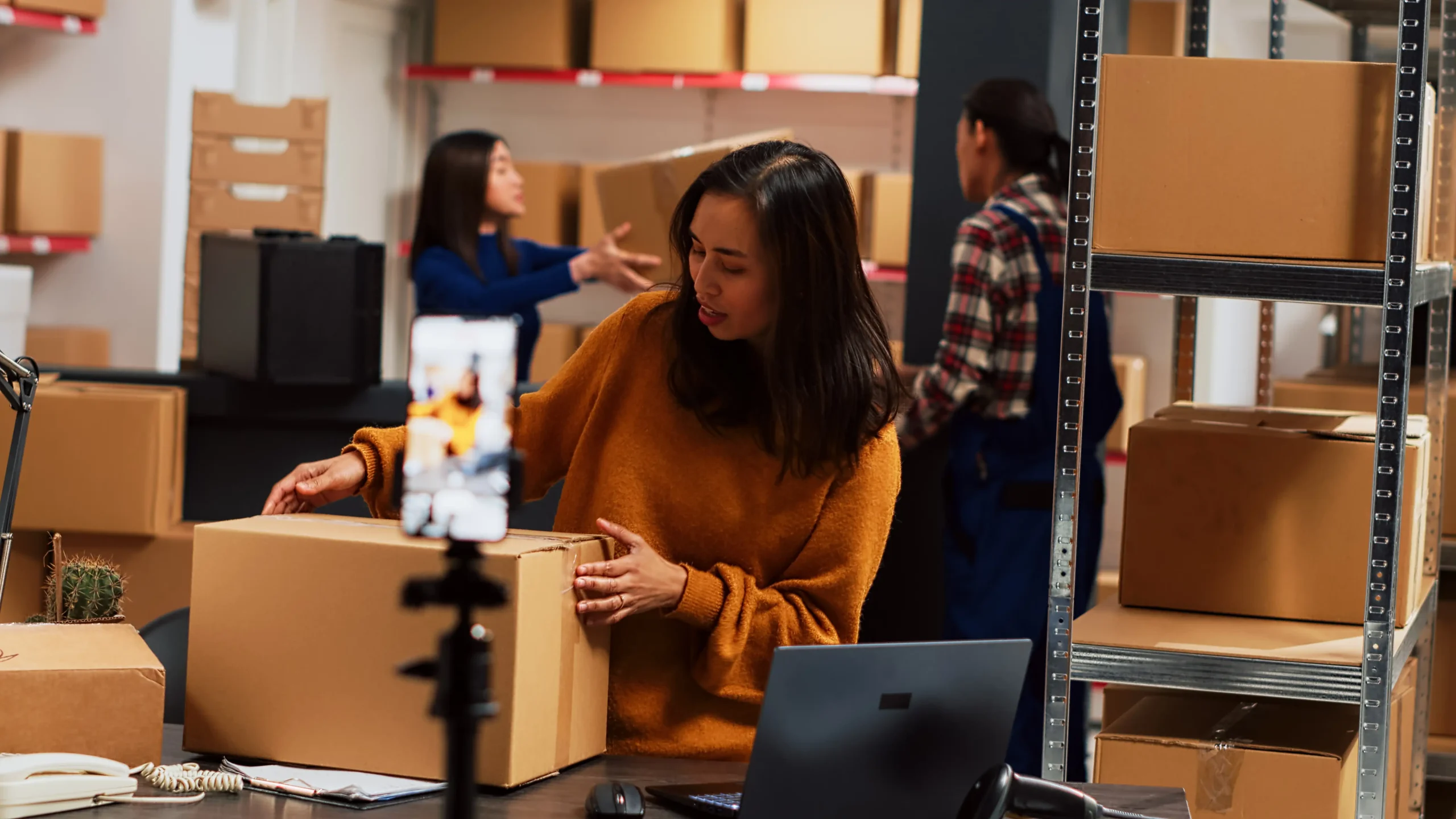 Warehouse employee preparing e-commerce packages while filming a marketing video with a smartphone.