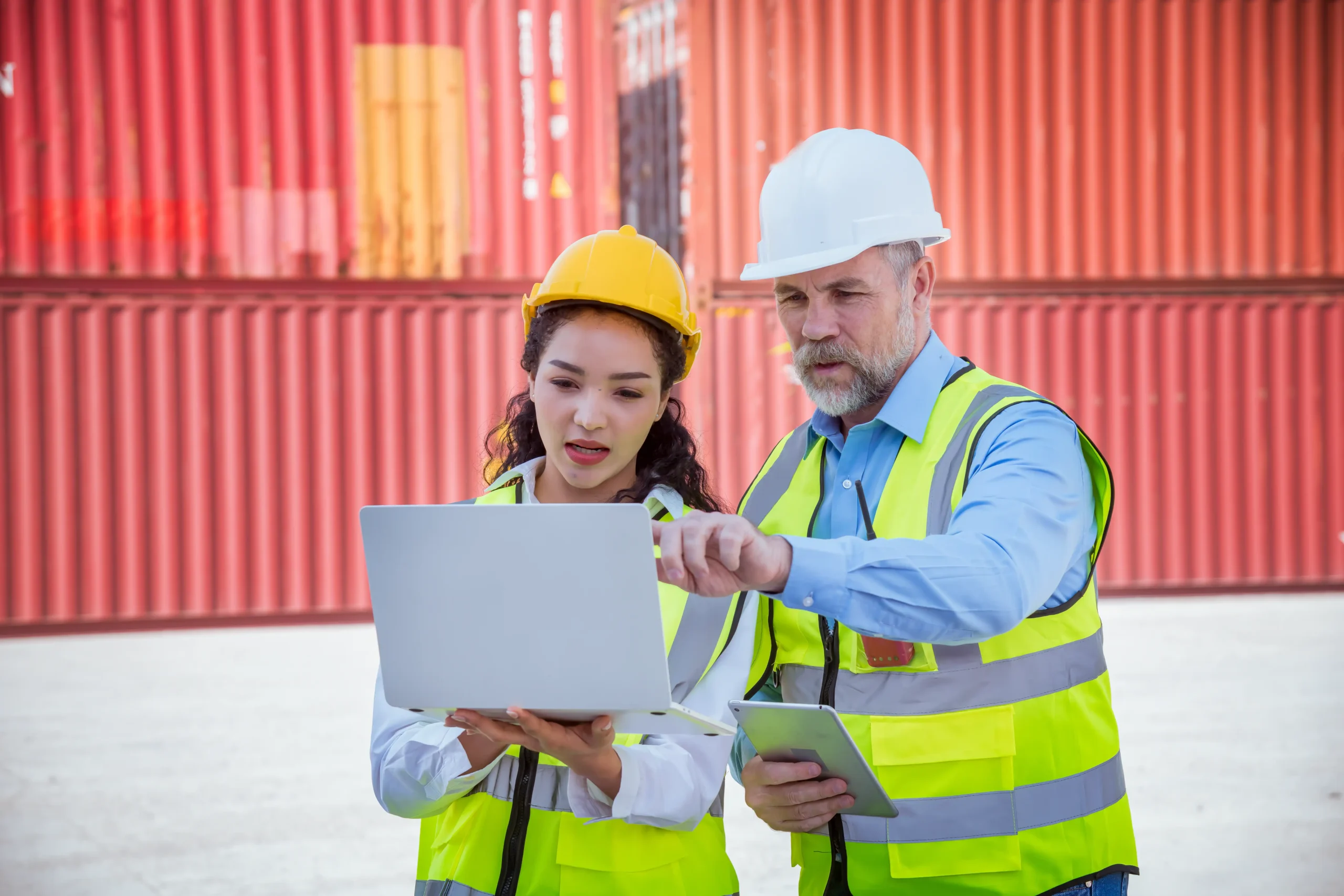 Dock workers discussing logistics software on a laptop in front of cargo containers.