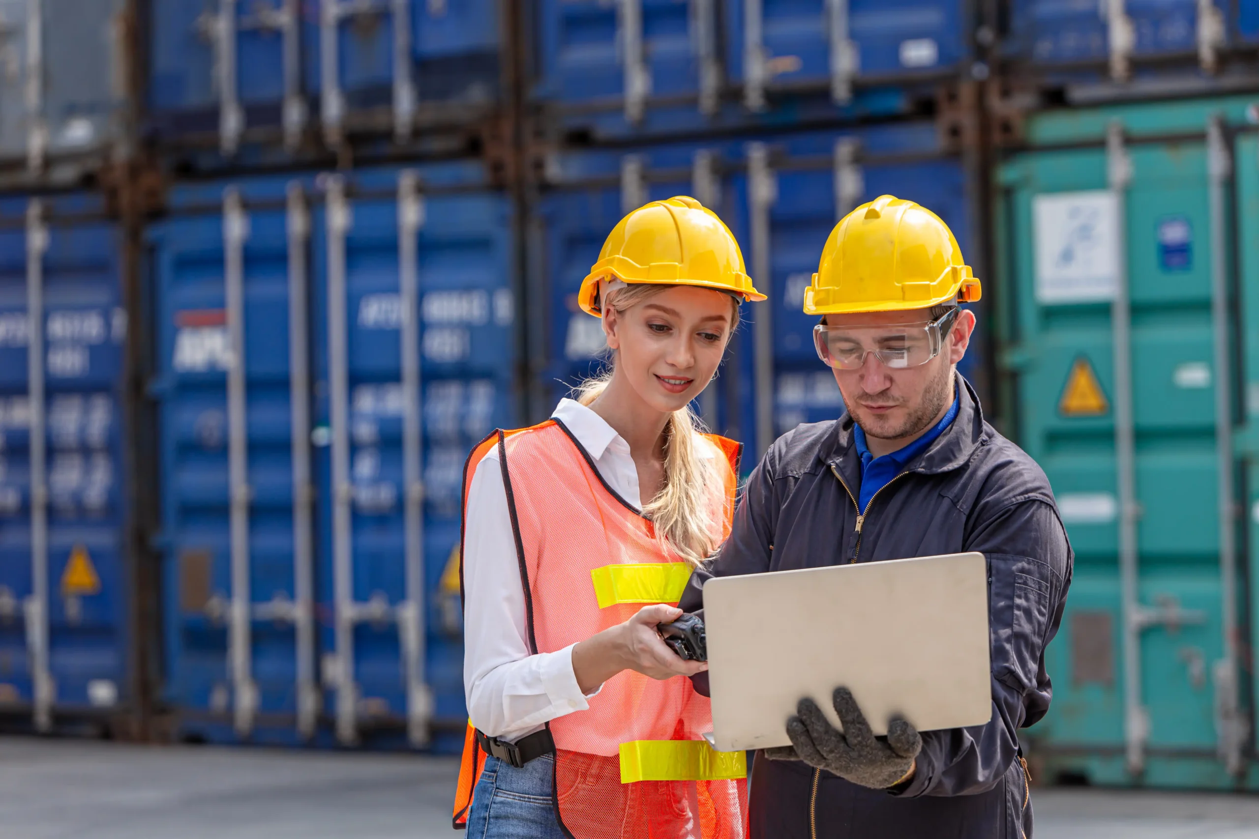 Port employees reviewing logistics software on a laptop with shipping containers in the background.