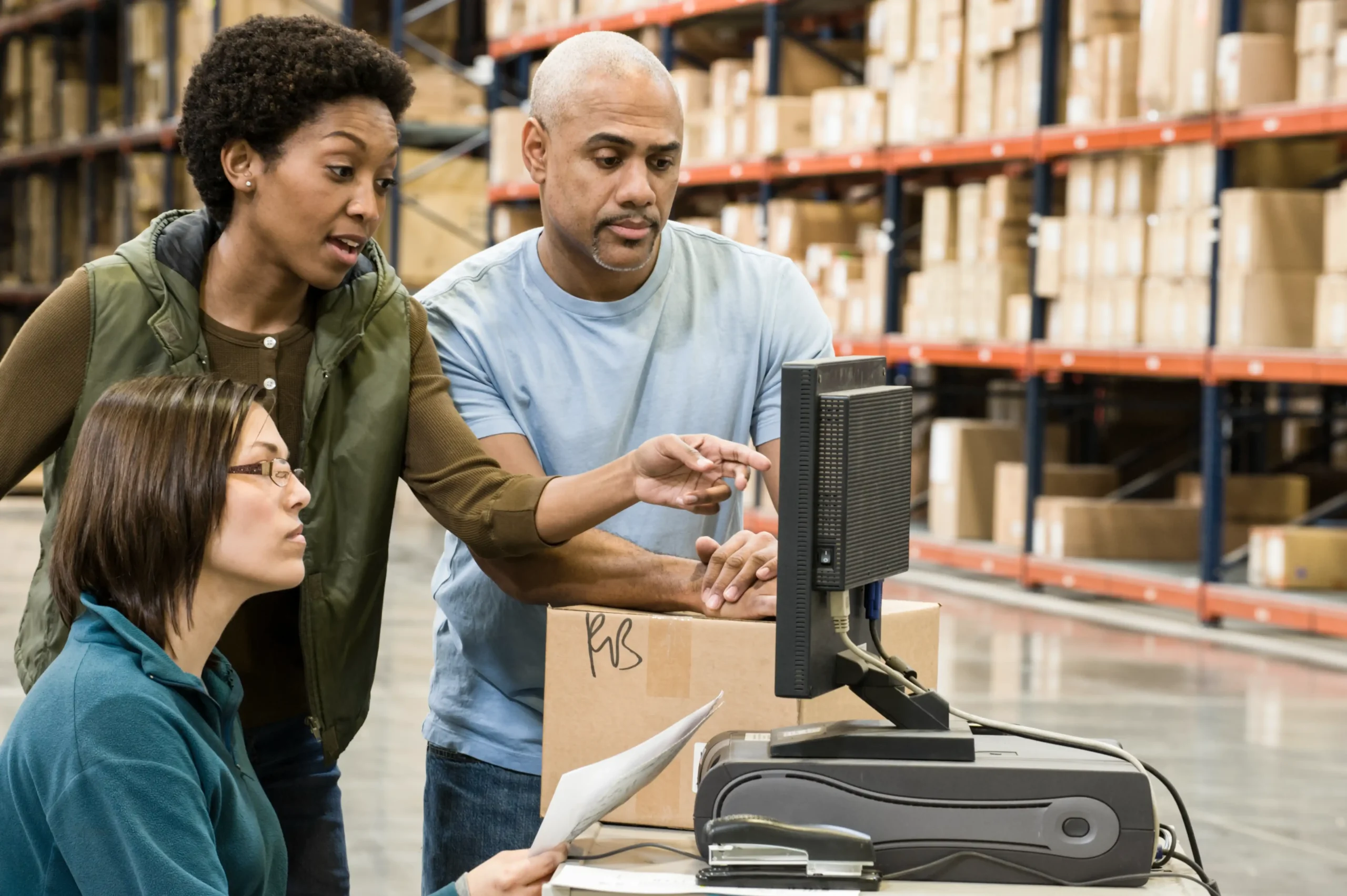 Warehouse employees collaborating on inventory tracking with logistics software on a desktop computer.