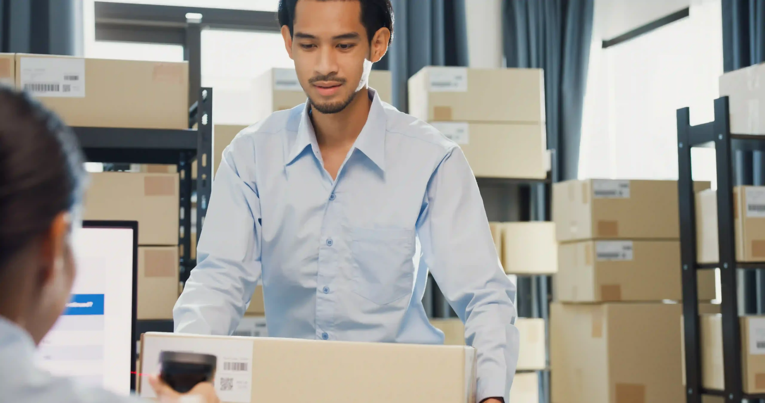 A warehouse worker scans and prepares a cardboard box for shipment in a fulfillment center, surrounded by inventory shelves.