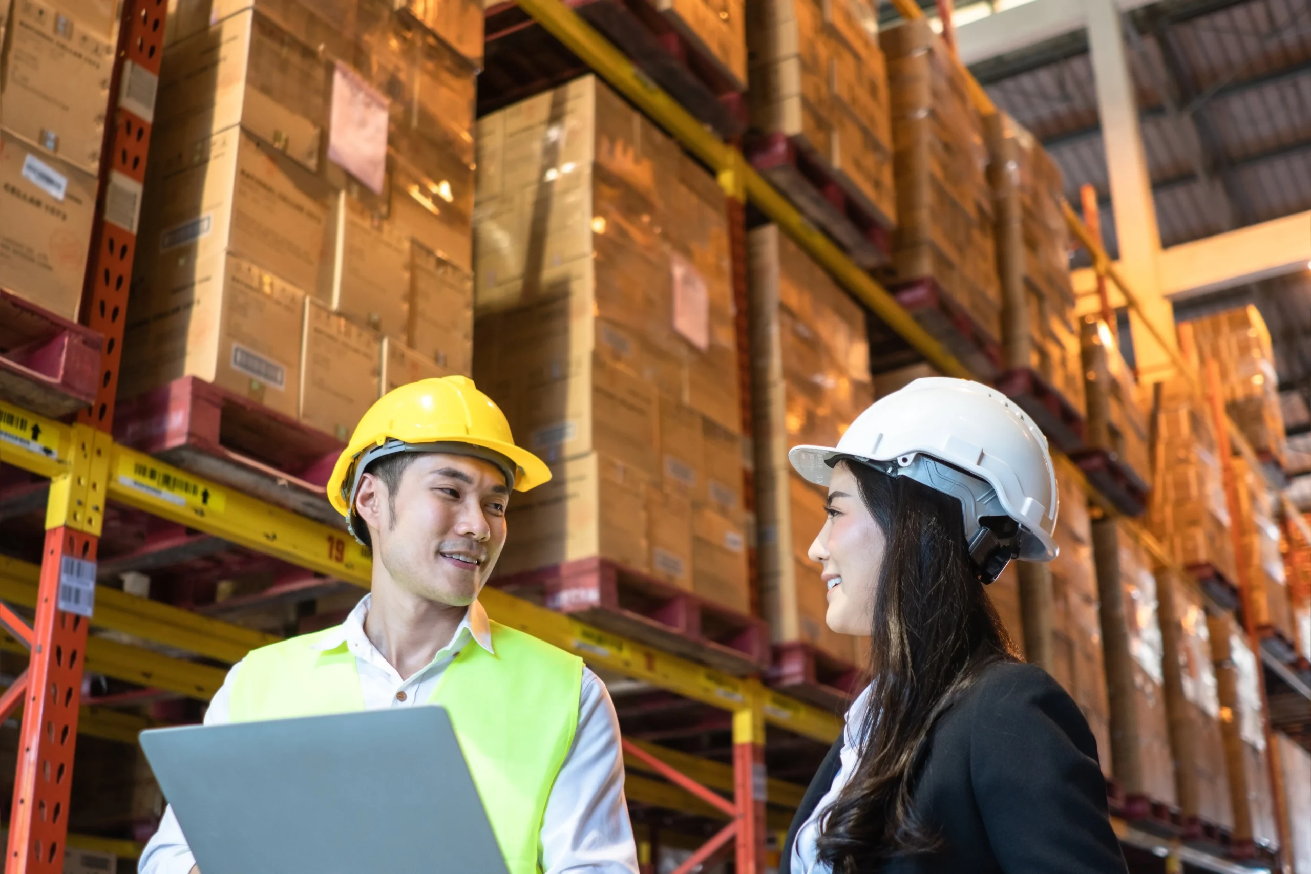 Asian male contractor discussing supply chain logistics with a female colleague in a warehouse surrounded by stacked goods.