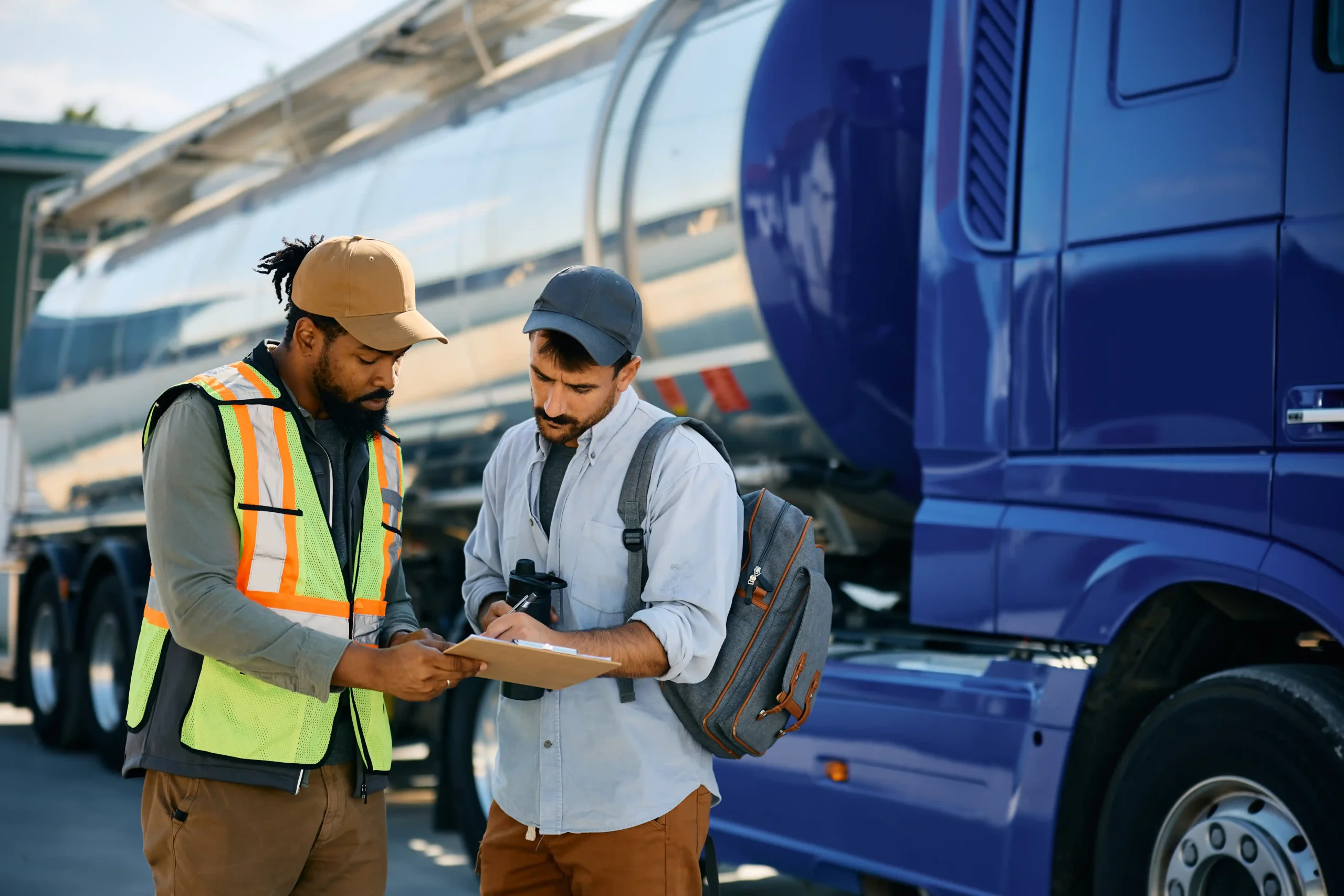 A black dispatcher and a truck driver discussing logistics near a large blue tanker truck.