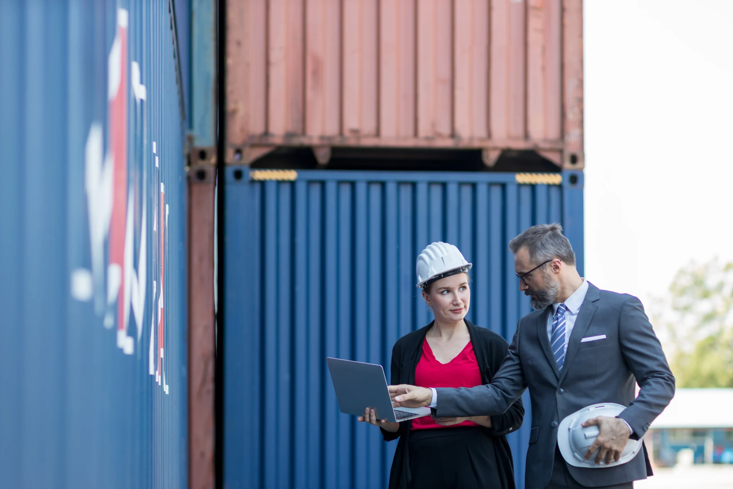 A businessman and a secretary in formal attire discussing logistics operations while standing near shipping containers.