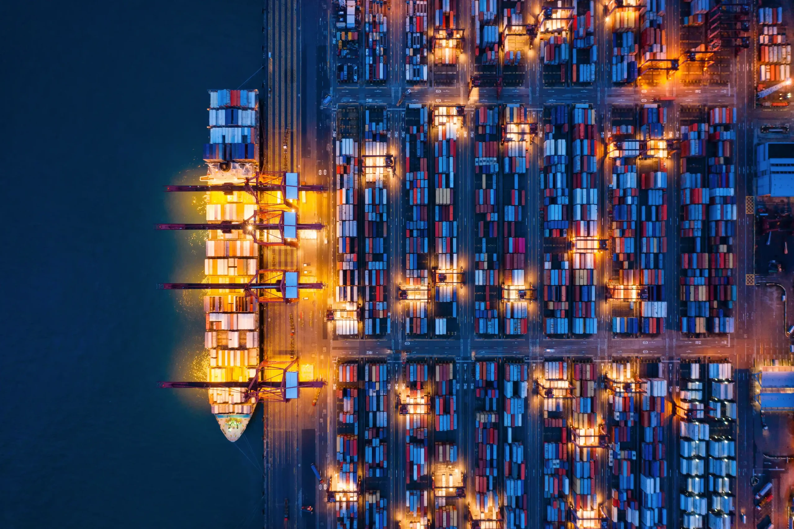 Aerial view of a container cargo ship and illuminated shipping docks during nighttime operations.