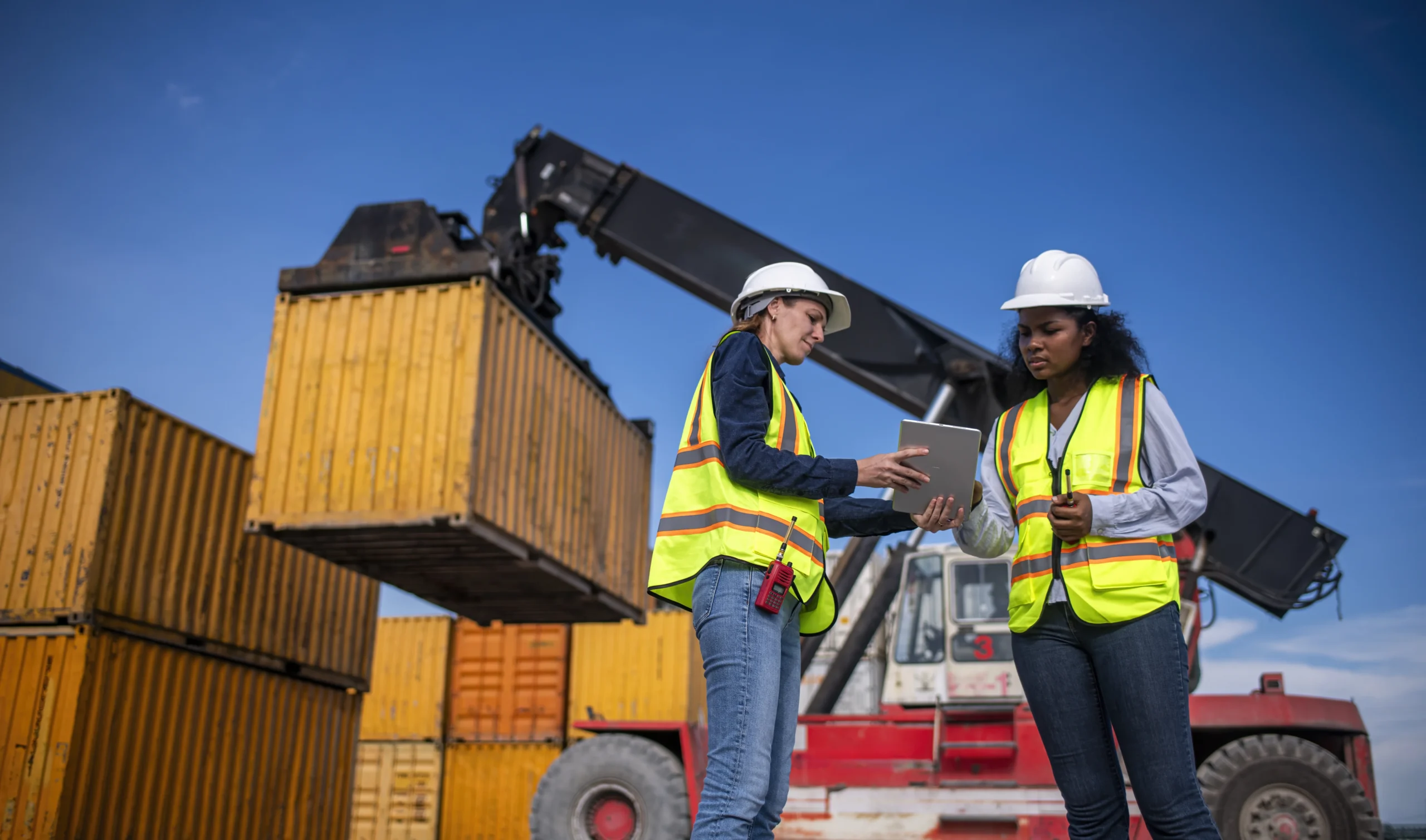 Two female dockworkers in safety vests and helmets reviewing a logistics plan on a tablet near a crane loading shipping containers.