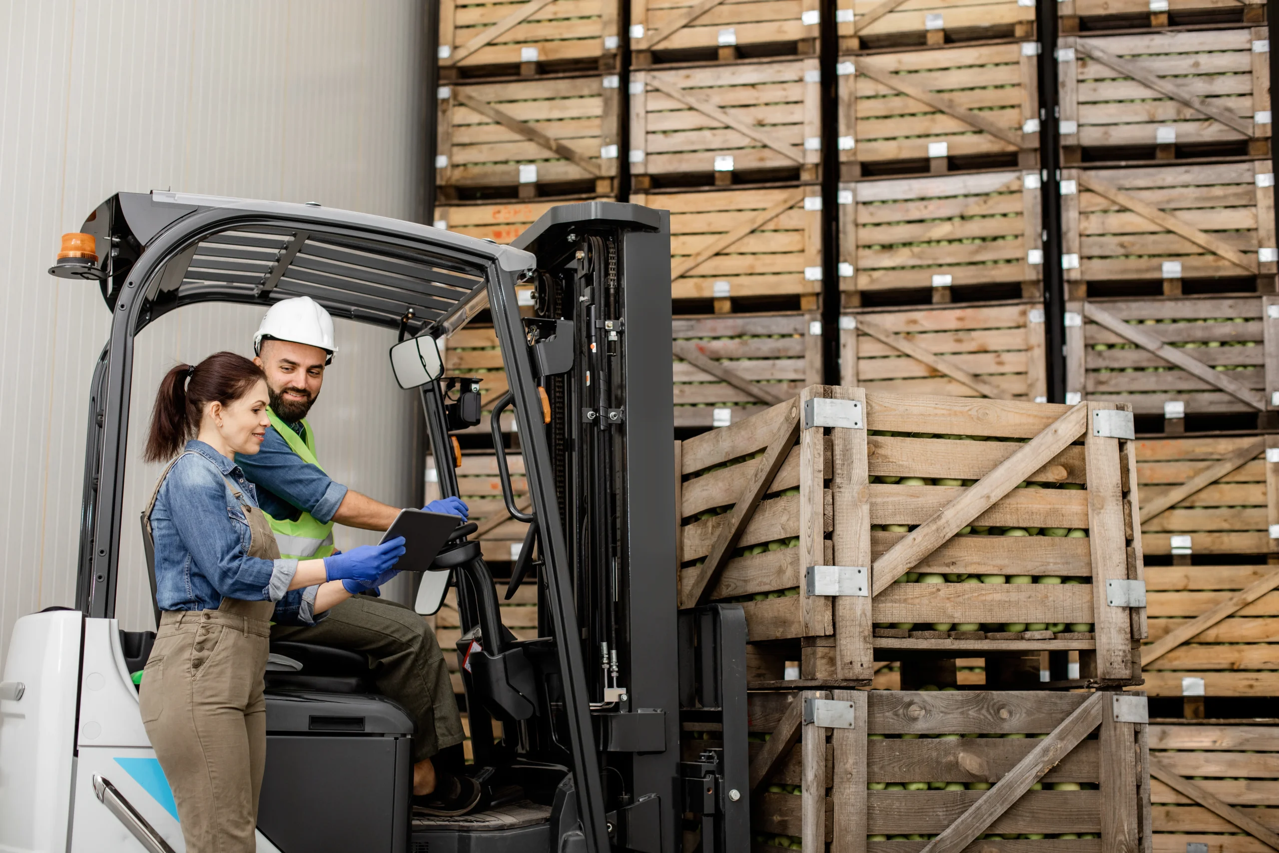 A forklift operator and a worker collaborating with a digital tablet in a warehouse with wooden pallets stacked in the background.