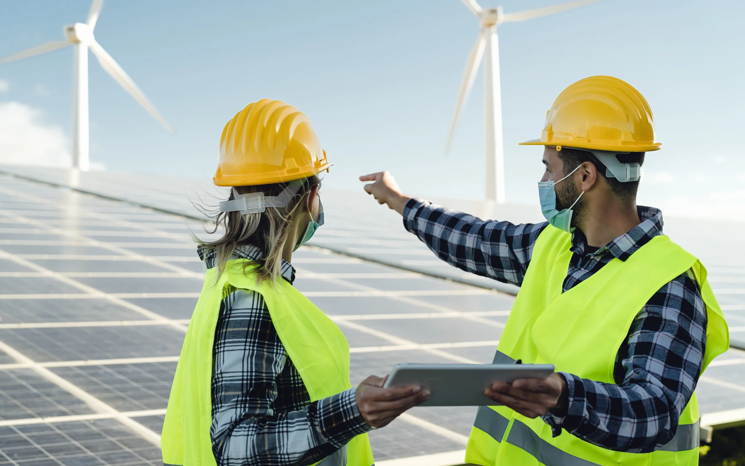 Two renewable energy engineers inspecting solar panels with wind turbines in the background.