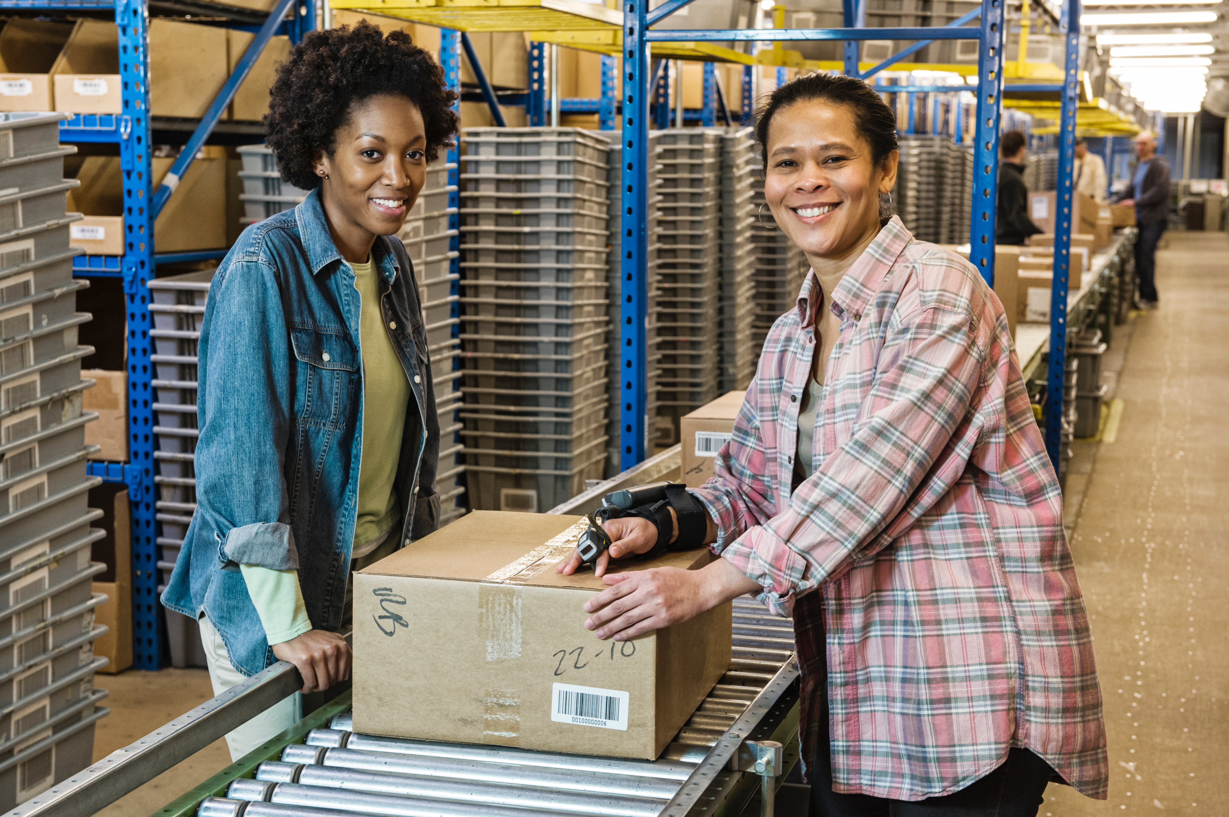 Two female warehouse workers smiling while organizing packages in a well-optimized warehouse storage facility with shelves and conveyors.