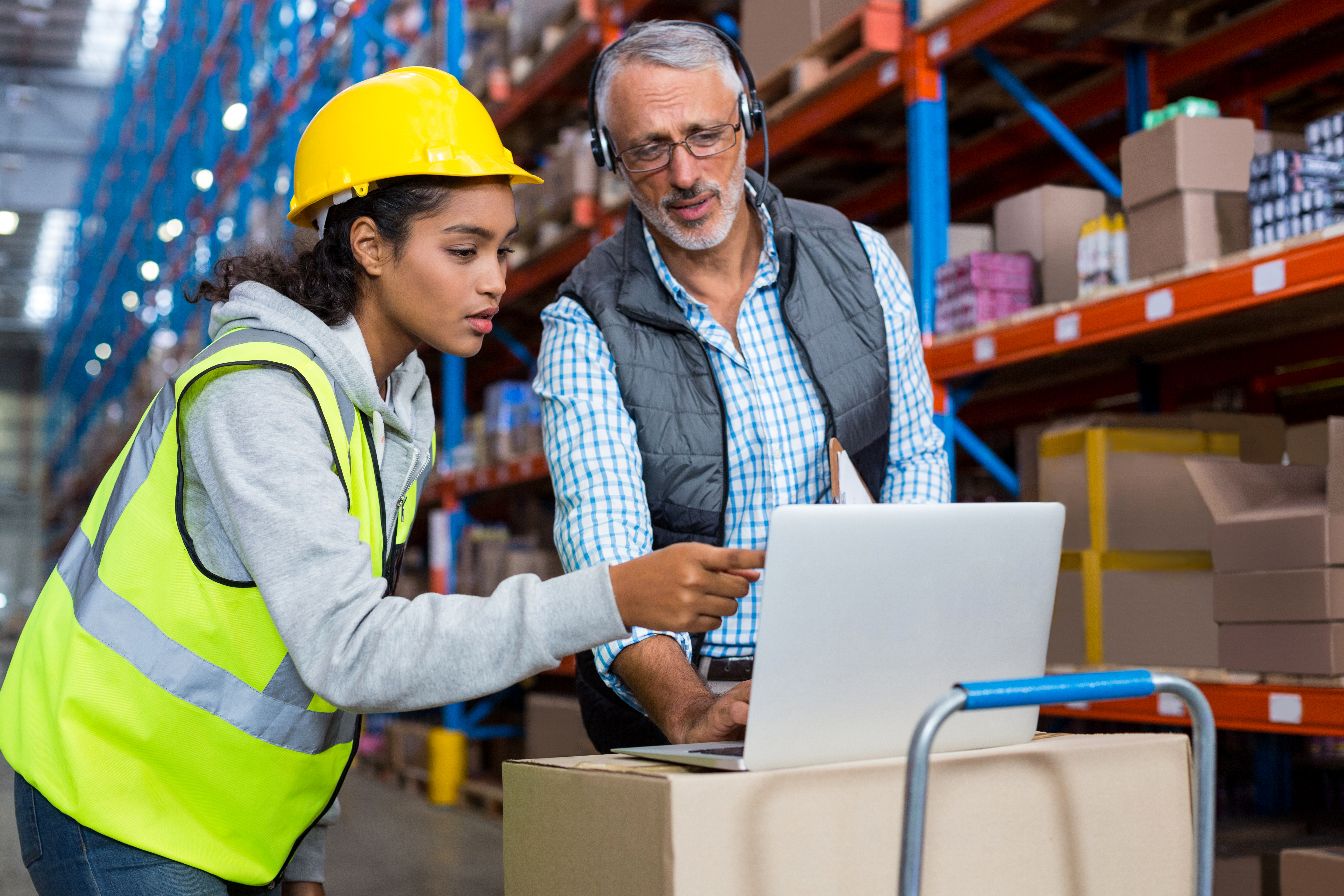 A warehouse manager and a worker in a safety vest and helmet collaborate on a laptop in a large warehouse, showcasing inventory management and operational efficiency.