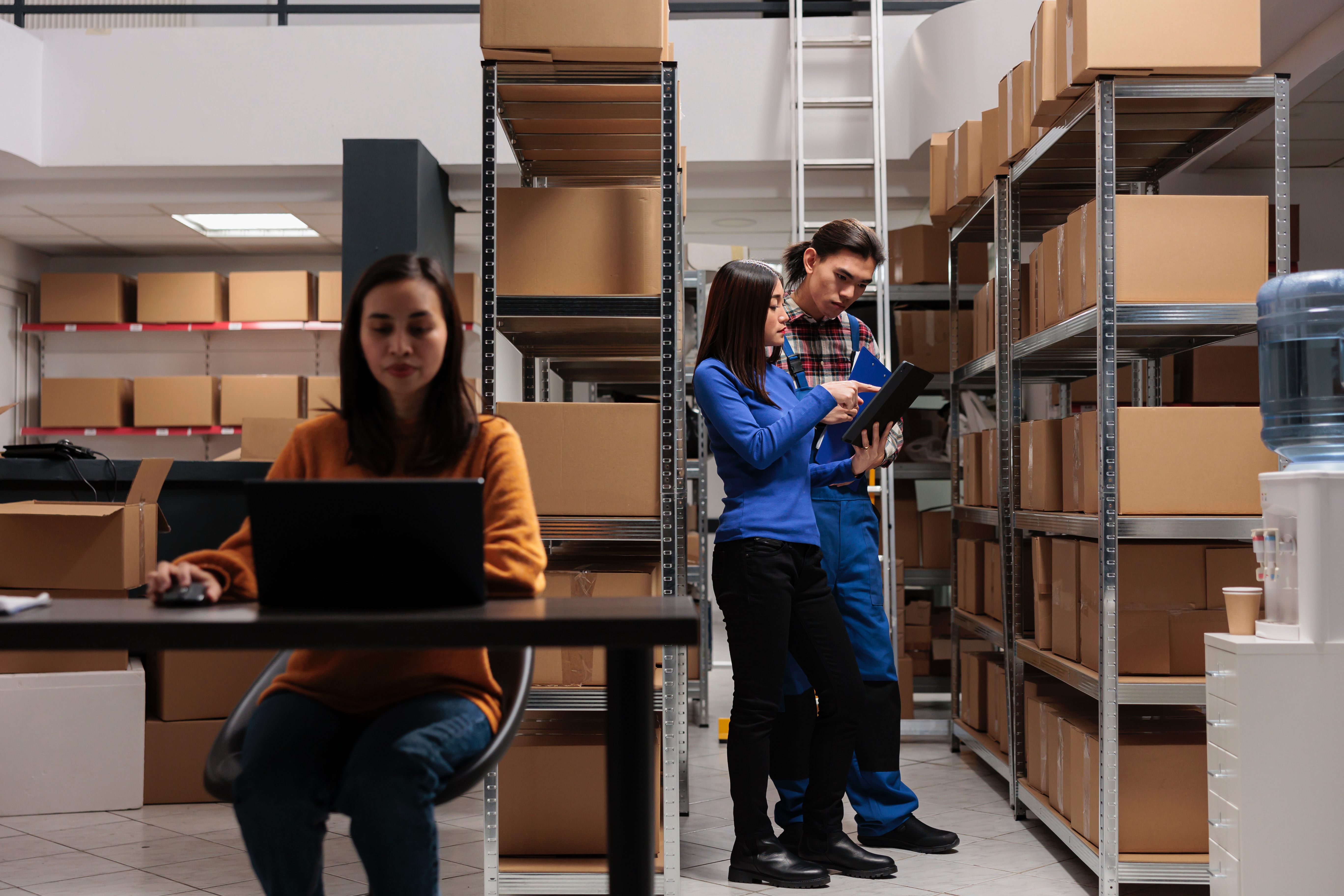 Warehouse team managing inventory with shelving systems, discussing solutions while another team member works on a laptop in the foreground.