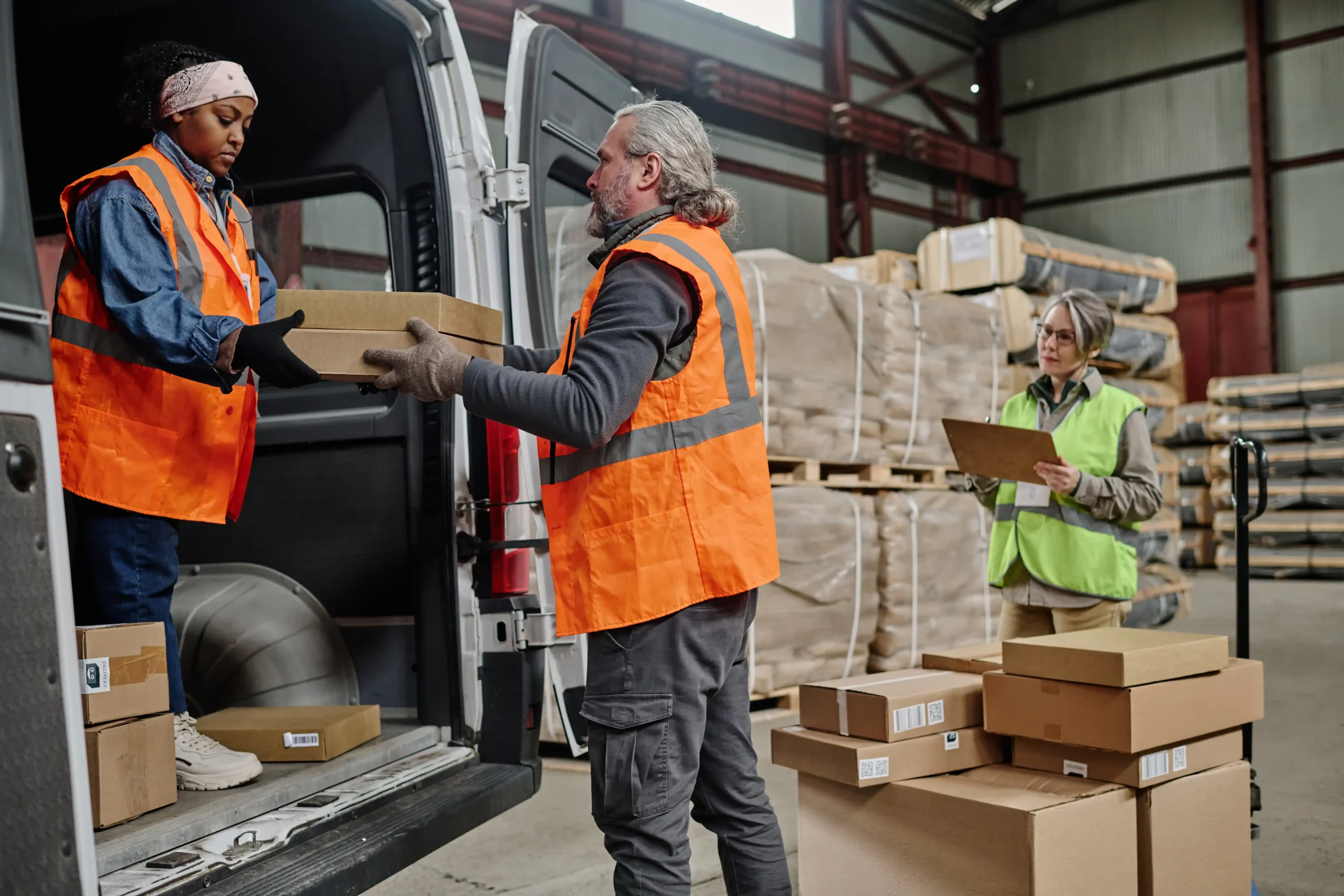 Warehouse workers collaborating to load packages into a delivery van, wearing safety vests.