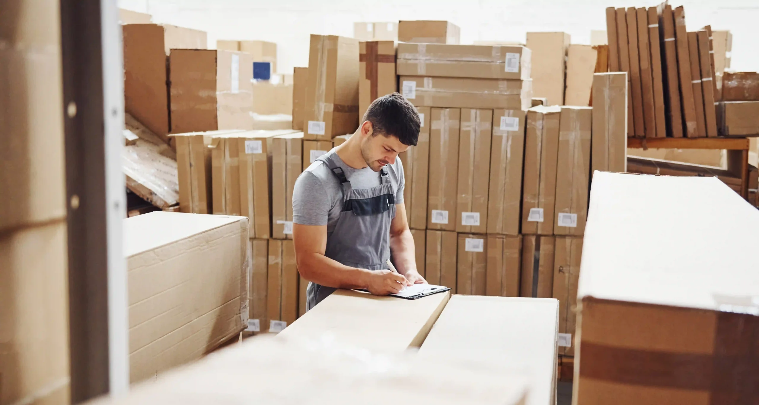 A warehouse worker in uniform reviews a checklist while surrounded by stacked cardboard boxes in a busy fulfillment center.