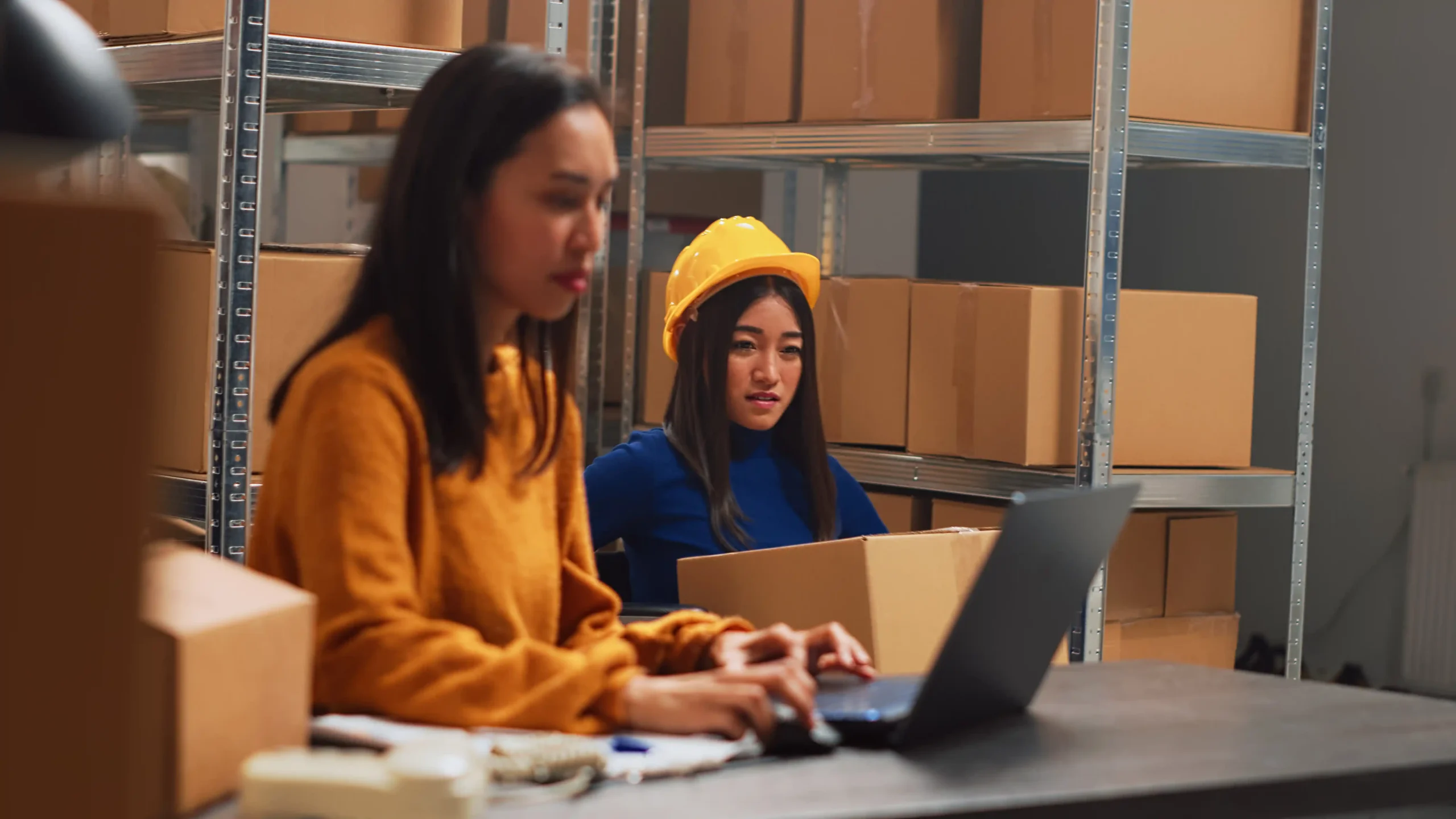 Two female workers in a supply chain warehouse collaborating on inventory and packing.