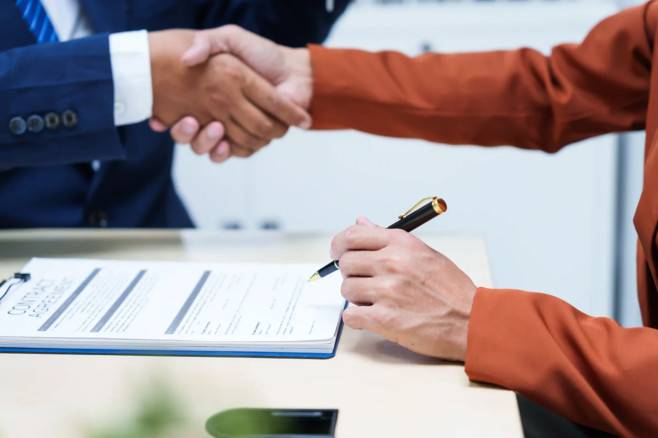 A close-up of a professional signing a logistics contract agreement at an office desk while shaking hands with a business partner.