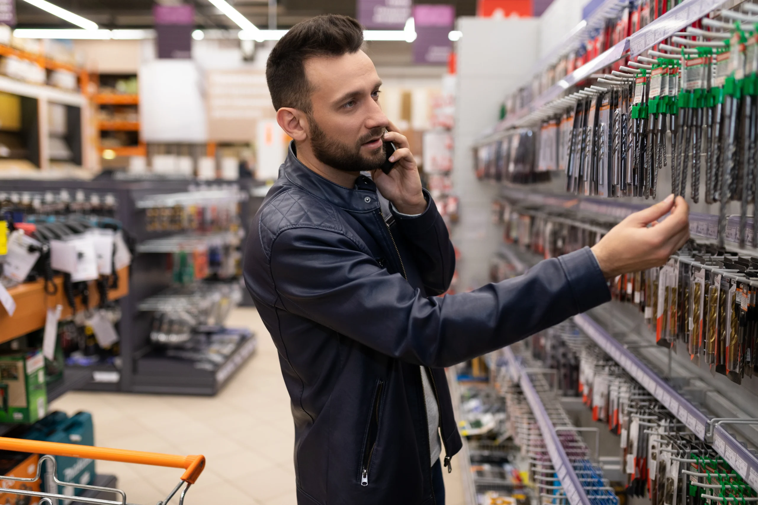 A male customer in a tool store browsing drill bits while talking on the phone, possibly reaching out to Pick and Save customer service for assistance.