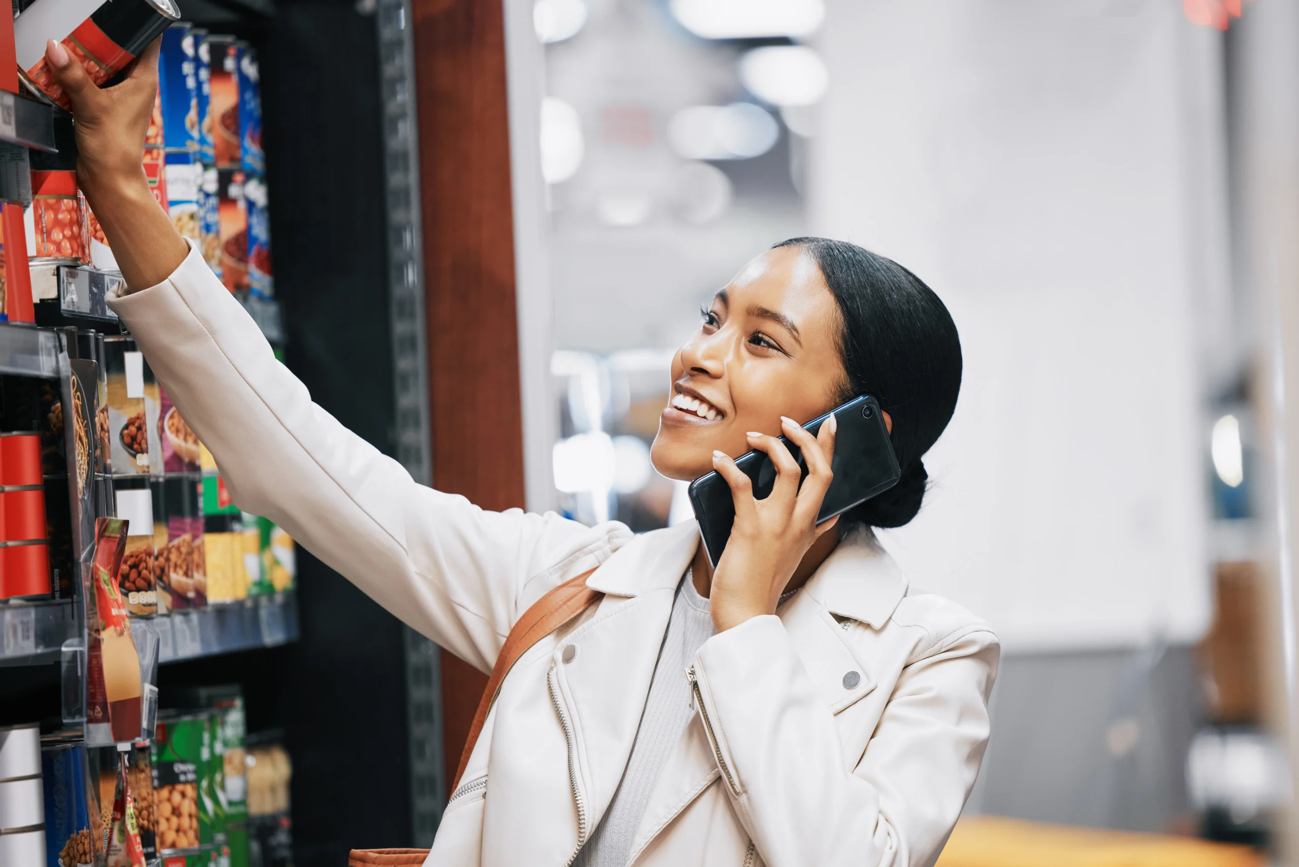 A happy woman in a supermarket aisle using her phone while selecting a product, possibly contacting Pick and Save customer service for product details.